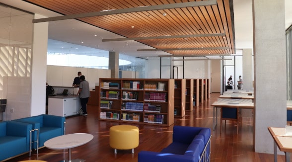 Uma biblioteca com sofás, banco, mesa e pessoas no fundo.